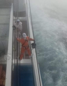 A ship is at sea. Two workers balance on the wires of a crane. They are at risk of falling into the sea.